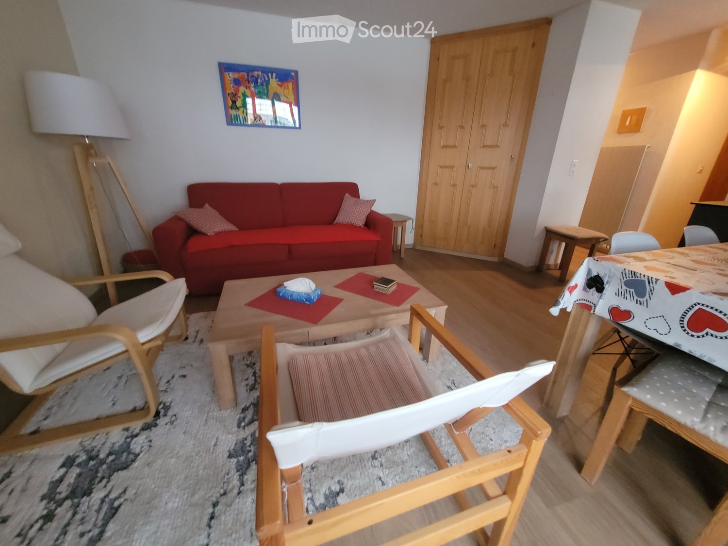 modern interior with hardwood floor, red couch with white pillows, wooden coffee table, wooden dining table, lamps, artwork on wall, rug