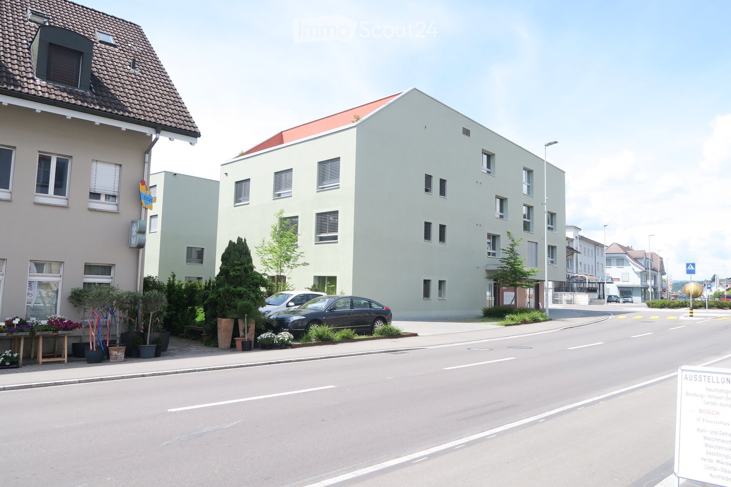 White modern building with red roof, several windows, two cars parked in front, greenery around the building