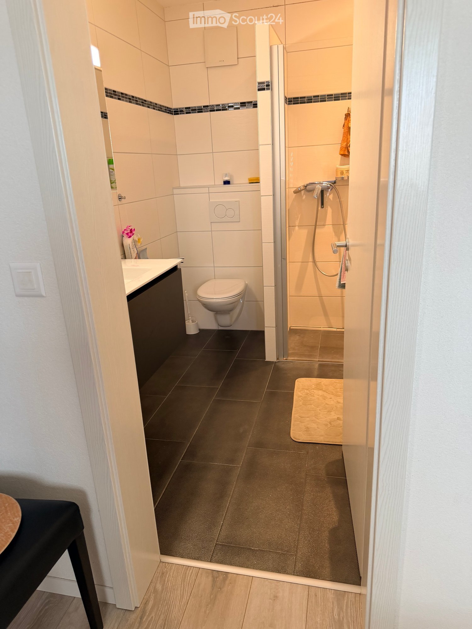 small bathroom with tile flooring, toilet, sink, shower stall, small bench, black wall cabinet, door to room