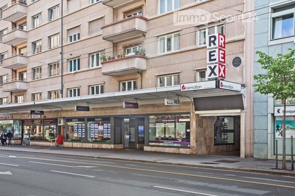 multi-story residential building, beige facade, multiple windows, balconies with potted plants, street-level commercial space, signage for 'REX', 'CLINIKX', 'BARNES', pedestrian walkway