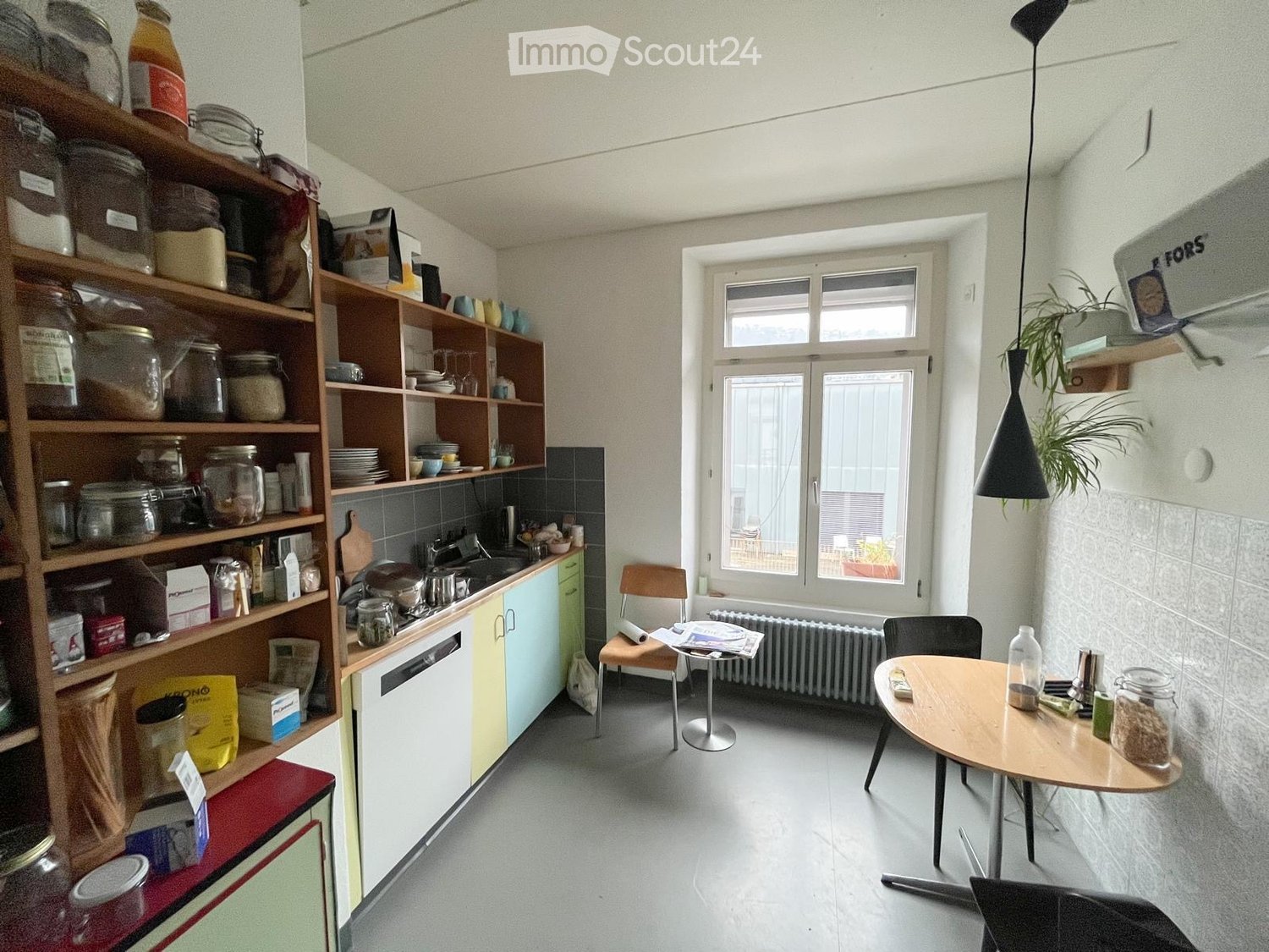 a small kitchen with a dining area, large shelves, window, radiator, table, chairs, and various kitchen items