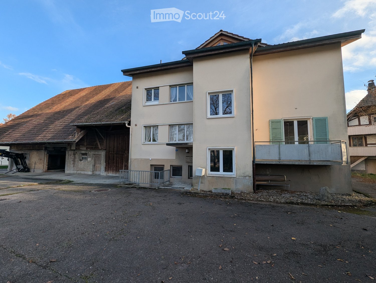 Two-story house with a barn, beige wall with windows, brown roof, metal railing on second floor balcony, and a paved driveway
