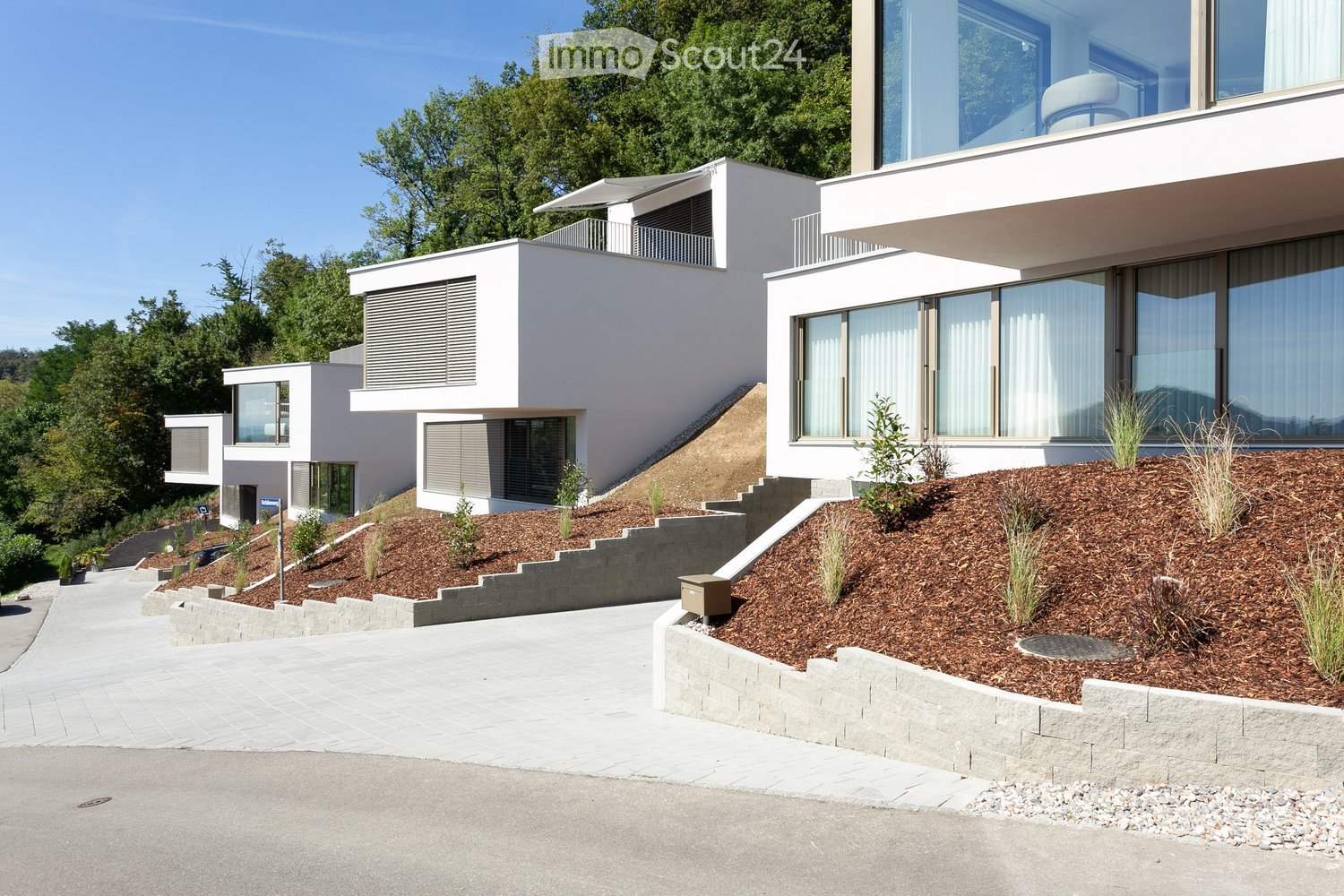 Modern apartment complex, white walls, large glass windows, raised ground floor, driveway, landscaped garden, mailboxes, distant treeline