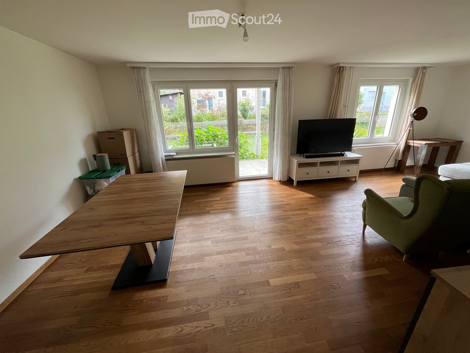 Empty room with wooden flooring, sliding glass door leading to patio, white television on TV stand, white radiators, green chair, dark wood table, and lamps