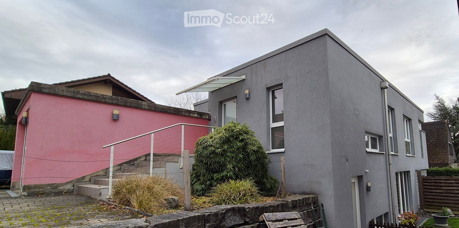 Modern house with flat roof, two different colored facades, a fenced front yard, stairs leading up to a raised ground floor.