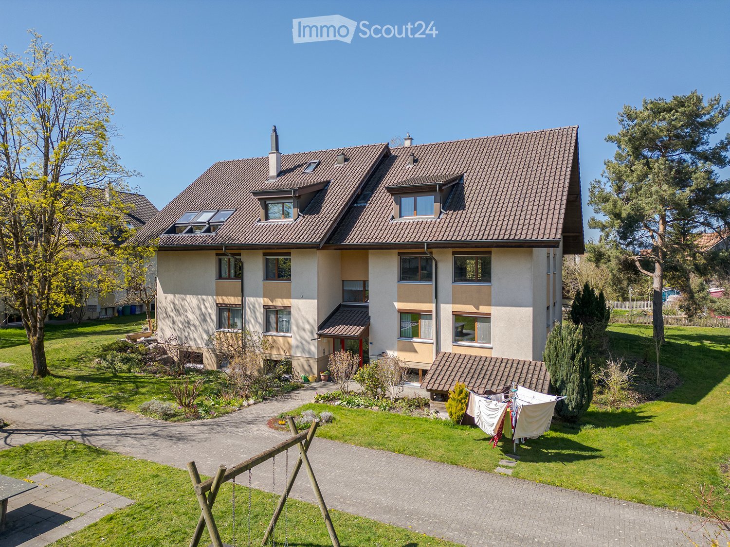 Two-story apartment building, brown roof, beige walls, several windows, driveway, yard with greenery, swing, clothes on clothesline.