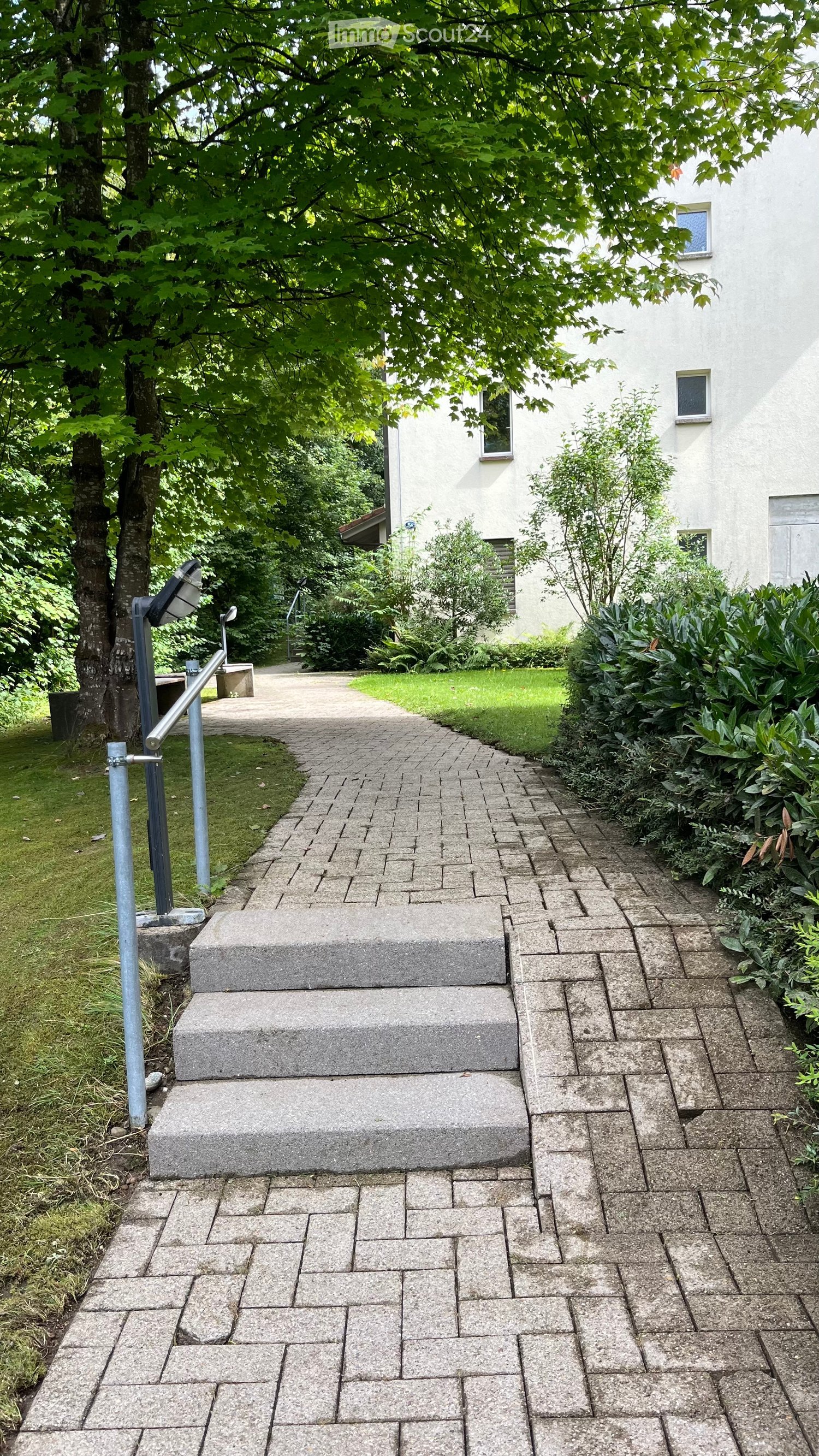 raised ground floor, concrete steps, paved pathway, tree, white building, bushes, metal handrail