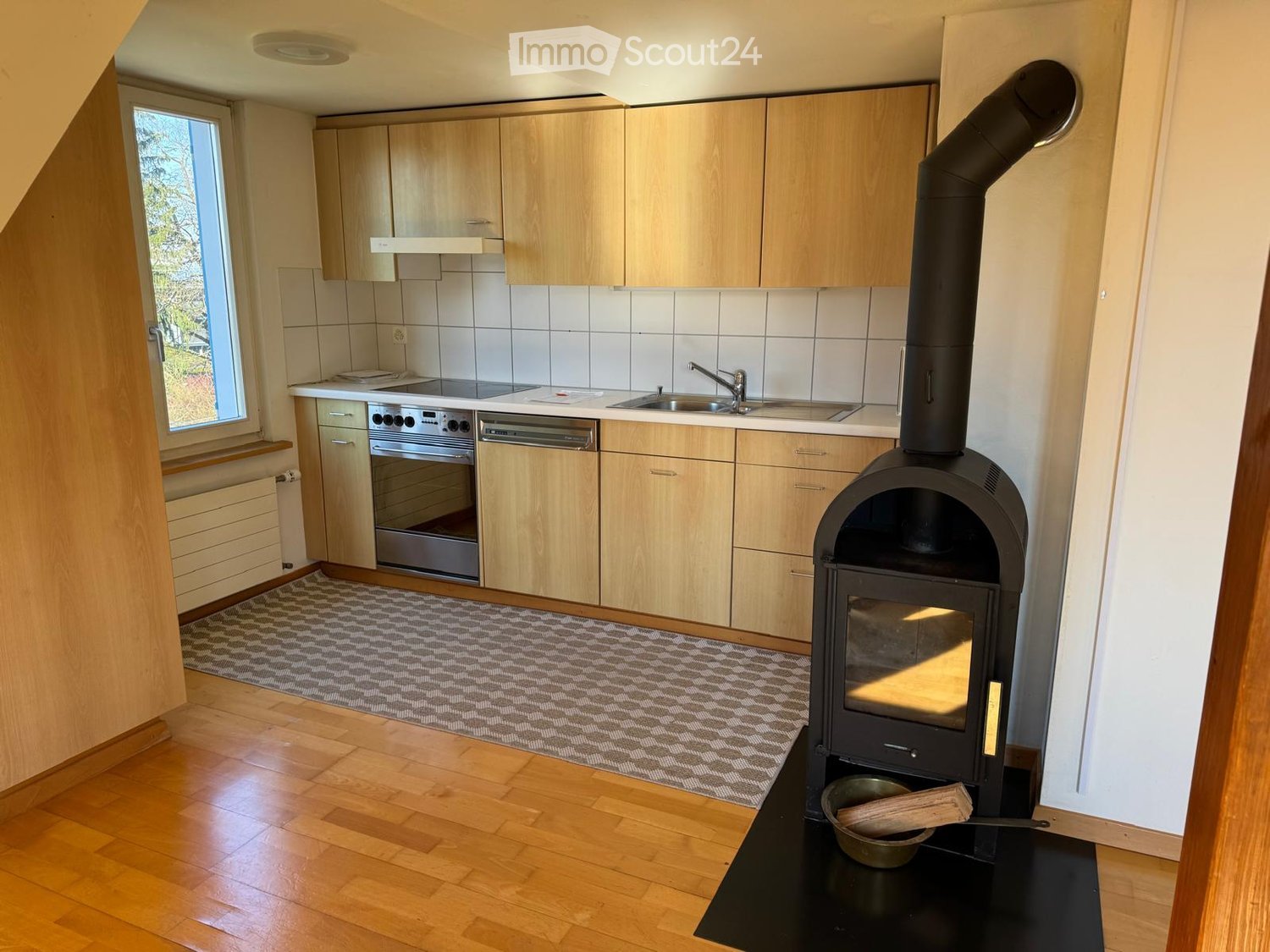 A kitchen with a gas stove, oven, sink, fireplace, wooden cabinets, tiled walls, and a rug.