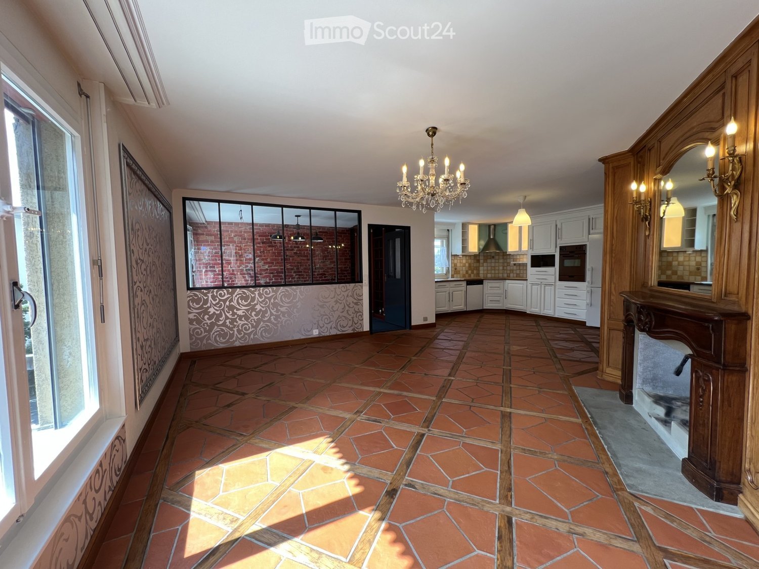 large open space, wooden tile floor, chandelier, fireplace, large window, kitchen in background