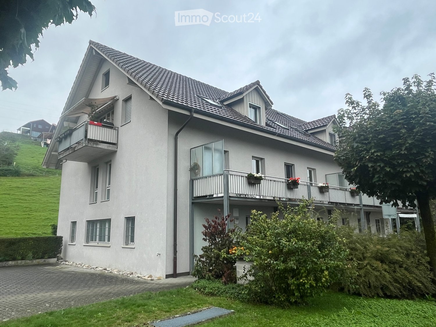 2-story house, gray walls, brown roof, two balconies with flowers, paved driveway