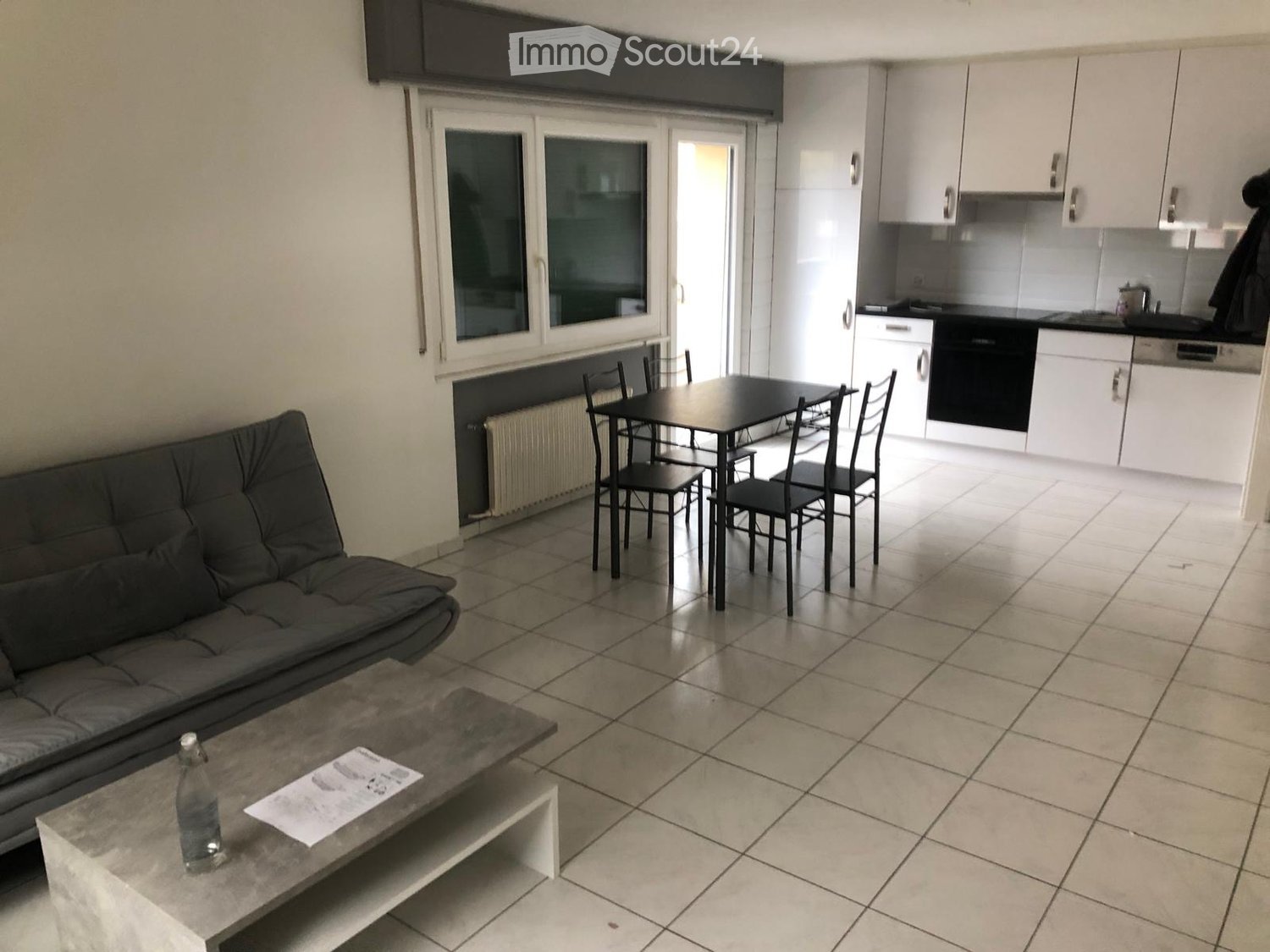 Empty living room, kitchen with dining area, white tiles, grey couch, central heating, integrated kitchen appliances