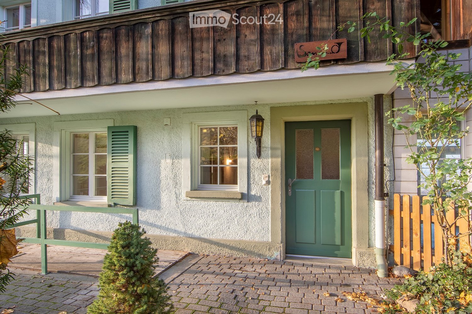 Facade with a wooden wall, 3 windows with shutters, an entrance with a green door, a lamp mounted on the wall.