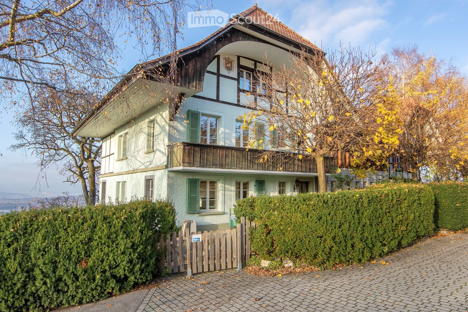 A two-story house with white walls and a brown roof, wooden balcony, hedges, and a small tree in front. It features a brown front door and several windows with shutters. A tree with yellow leaves is visible in front.