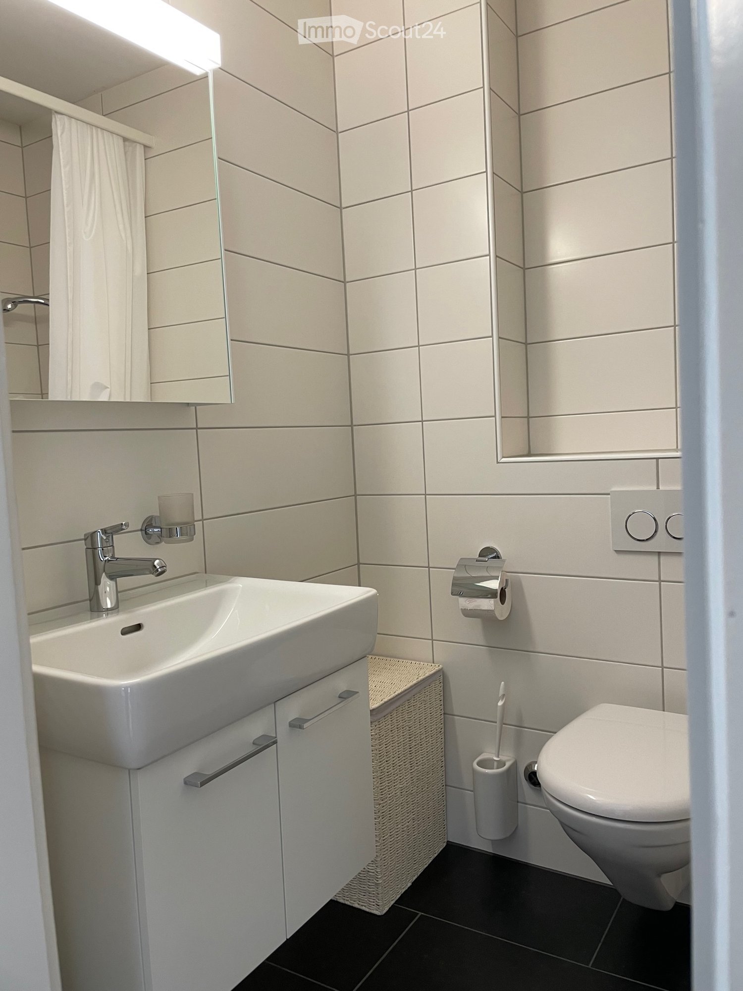 white tiled walls, black floor tiles, white sink with silver faucet, mirror, white toilet, white toilet paper holder, small wicker storage basket