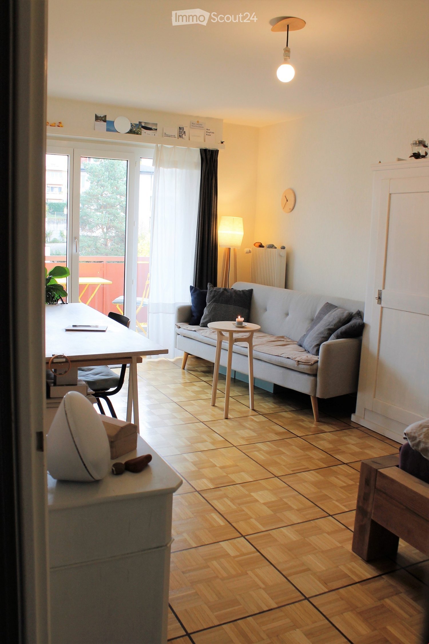 Living room with gray sofa, wooden side table, chair, desk, wooden flooring, radiator, lamp, white curtains, wall clock, and balcony access