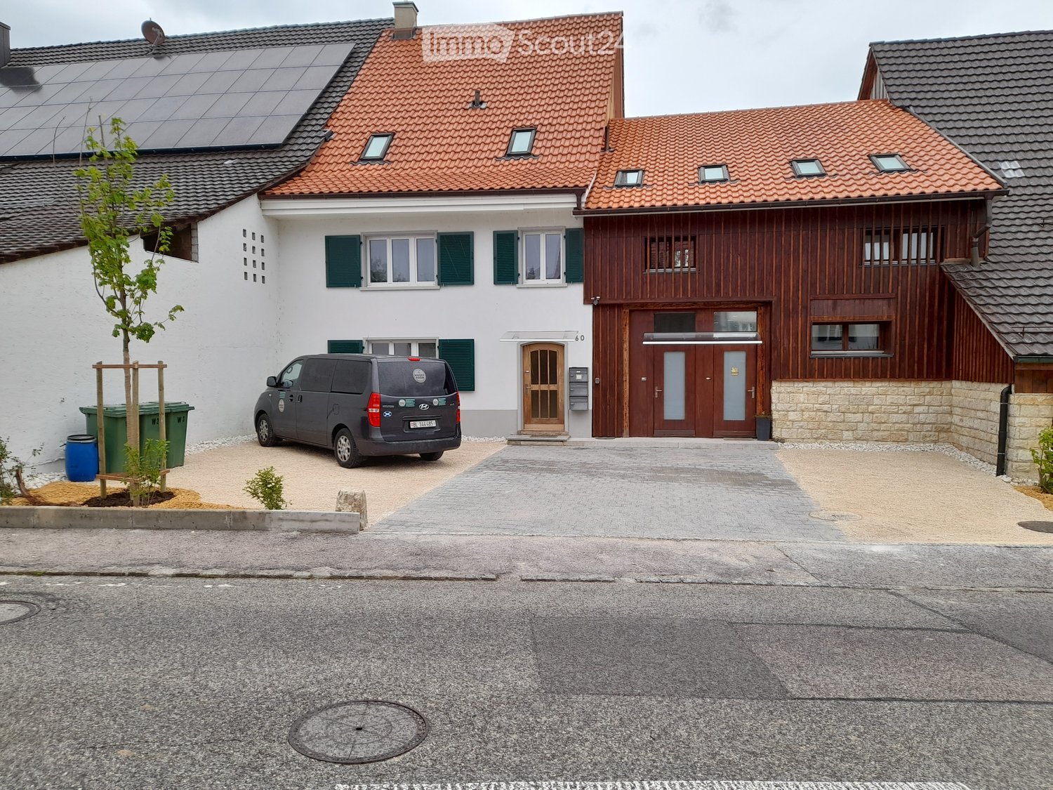 Two-story house, with a red tiled roof and green shutters, solar panels on the roof, gray van parked in front, wooden building attached to it, brick wall, and trash bins