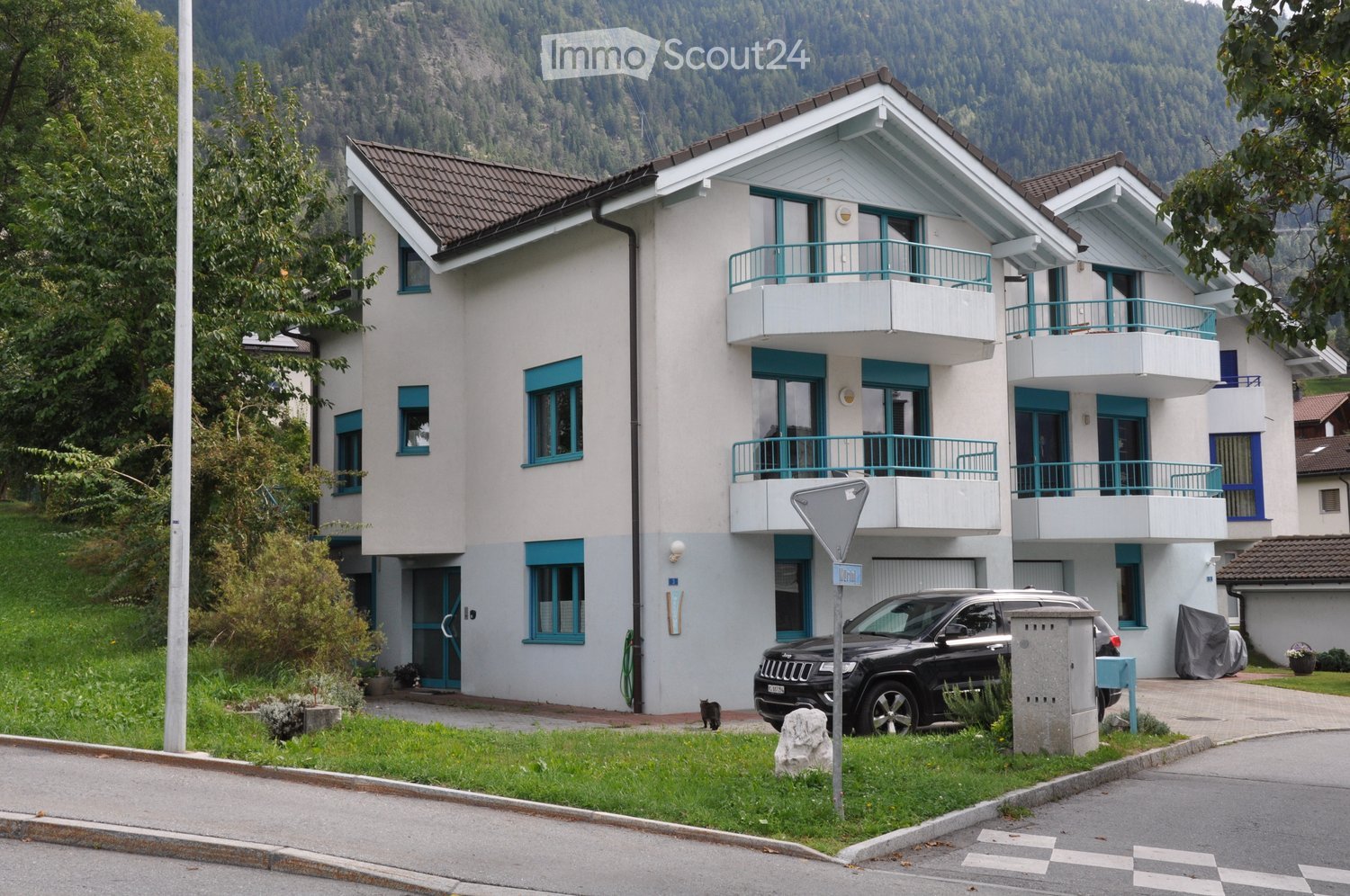 White two-story house with blue trim, balconies on the upper floor, located in a scenic area with mountains in the background, car parked in front