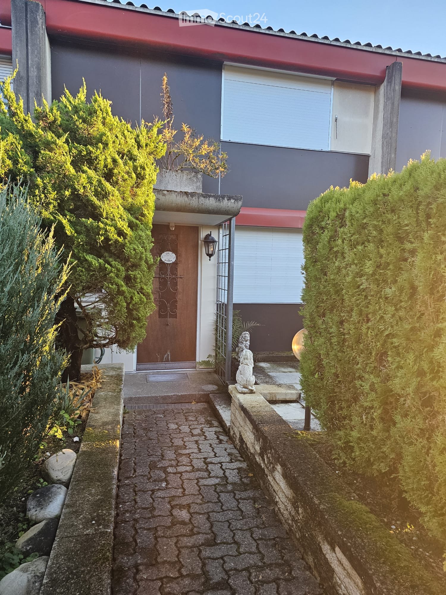 Entrance path with brick flooring, metal gate, wooden door, lamp, small fountain, shrub bushes