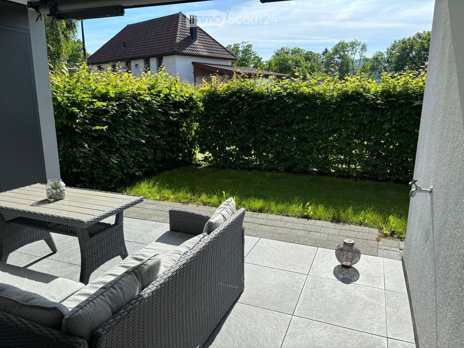 a seating area with a wicker sofa and table, paved terrace, a hedge in the background, and a grassy lawn