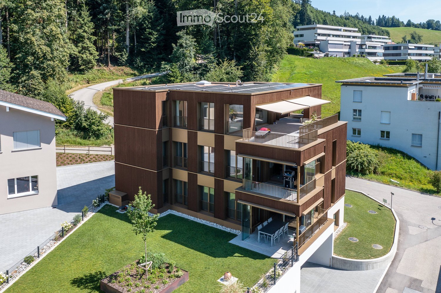Multi-story apartment building with balconies, solar panels on the roof, and a fenced grassy area.