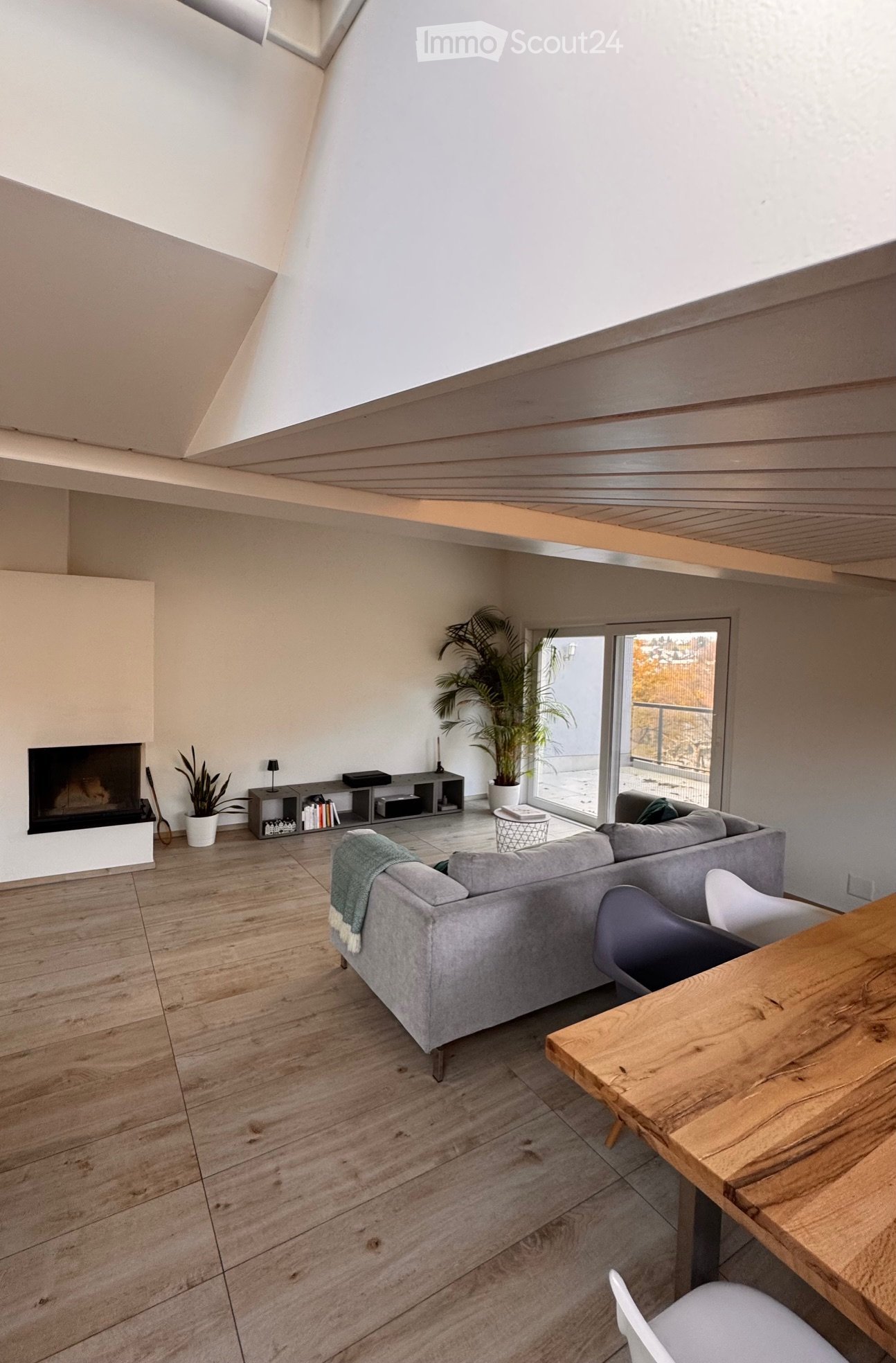 Open-plan living area, white walls, fireplace, wooden flooring, couch, wooden table, chairs, windows, plants