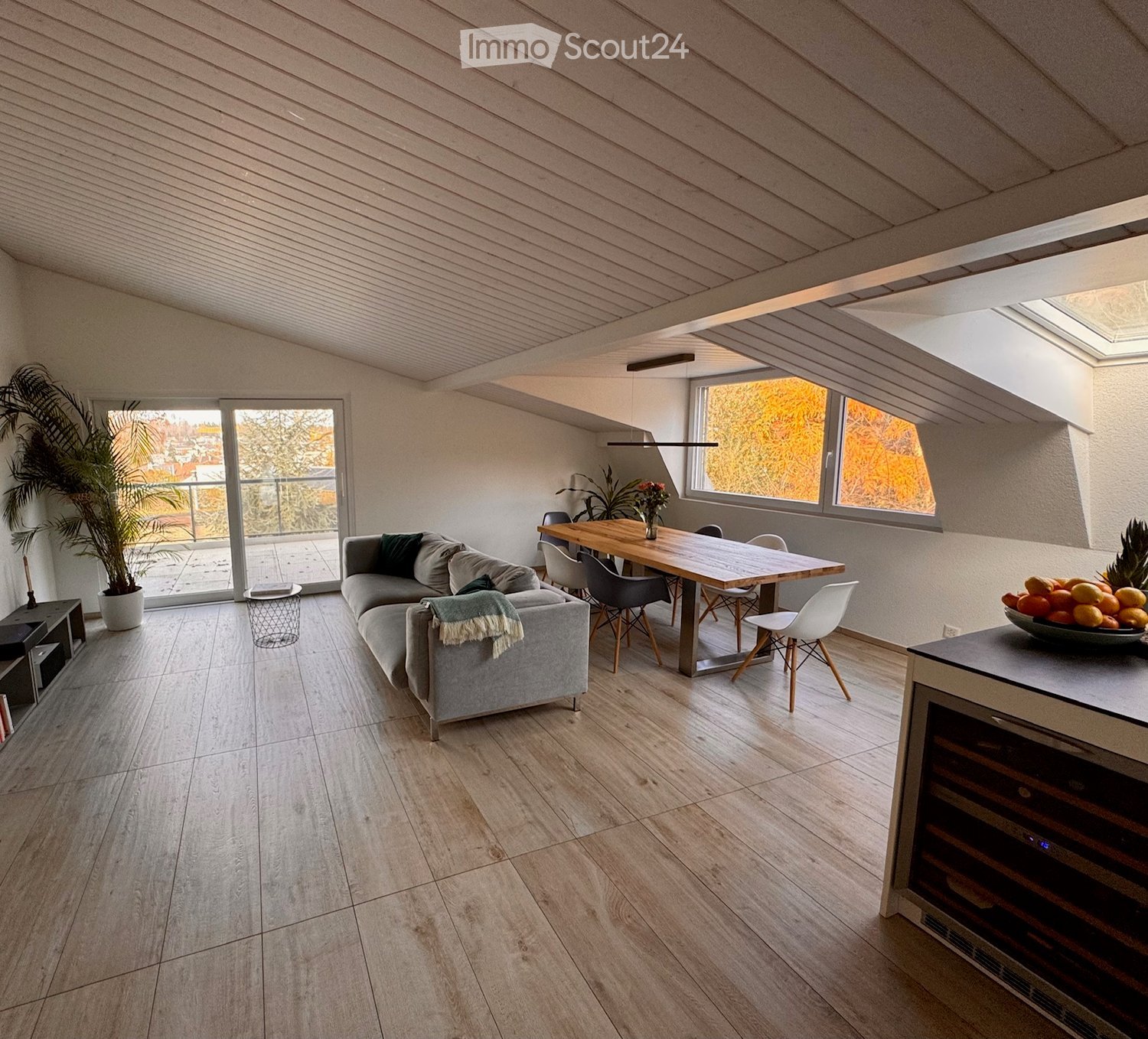 open plan, hardwood floors, dining table with chairs, gray sofa with pillows, side table, large sliding doors, balcony, windows, wooden table with flowers, fruit bowl on counter