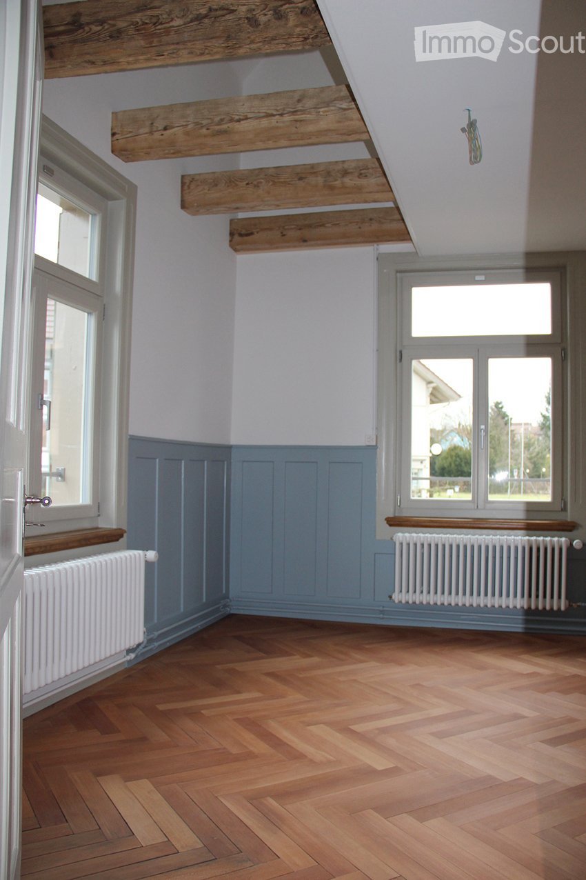 Wooden floors, white walls, blue wooden paneling, two windows, and white radiators.