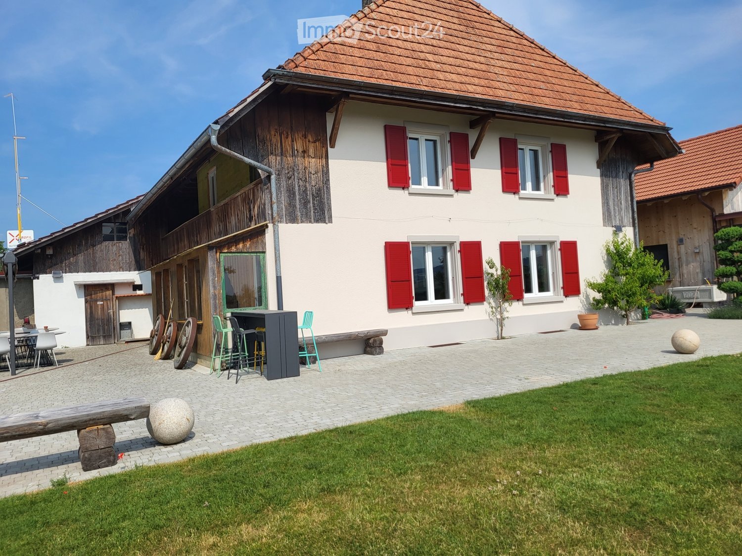 Two-story house with red shutters, white walls, brown roof, brick front, green lawn, outdoor seating area, wooden bench, outdoor table and chairs, potted plants, and a small fountain.