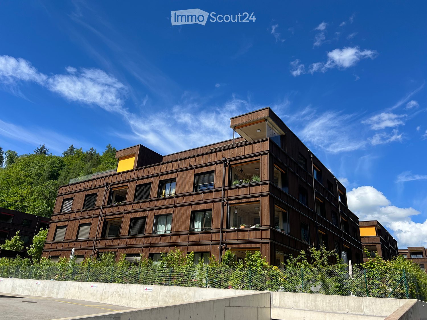 modern multi-storey building, brown wooden panels, glass windows, yellow accents, balconies, greenery in front