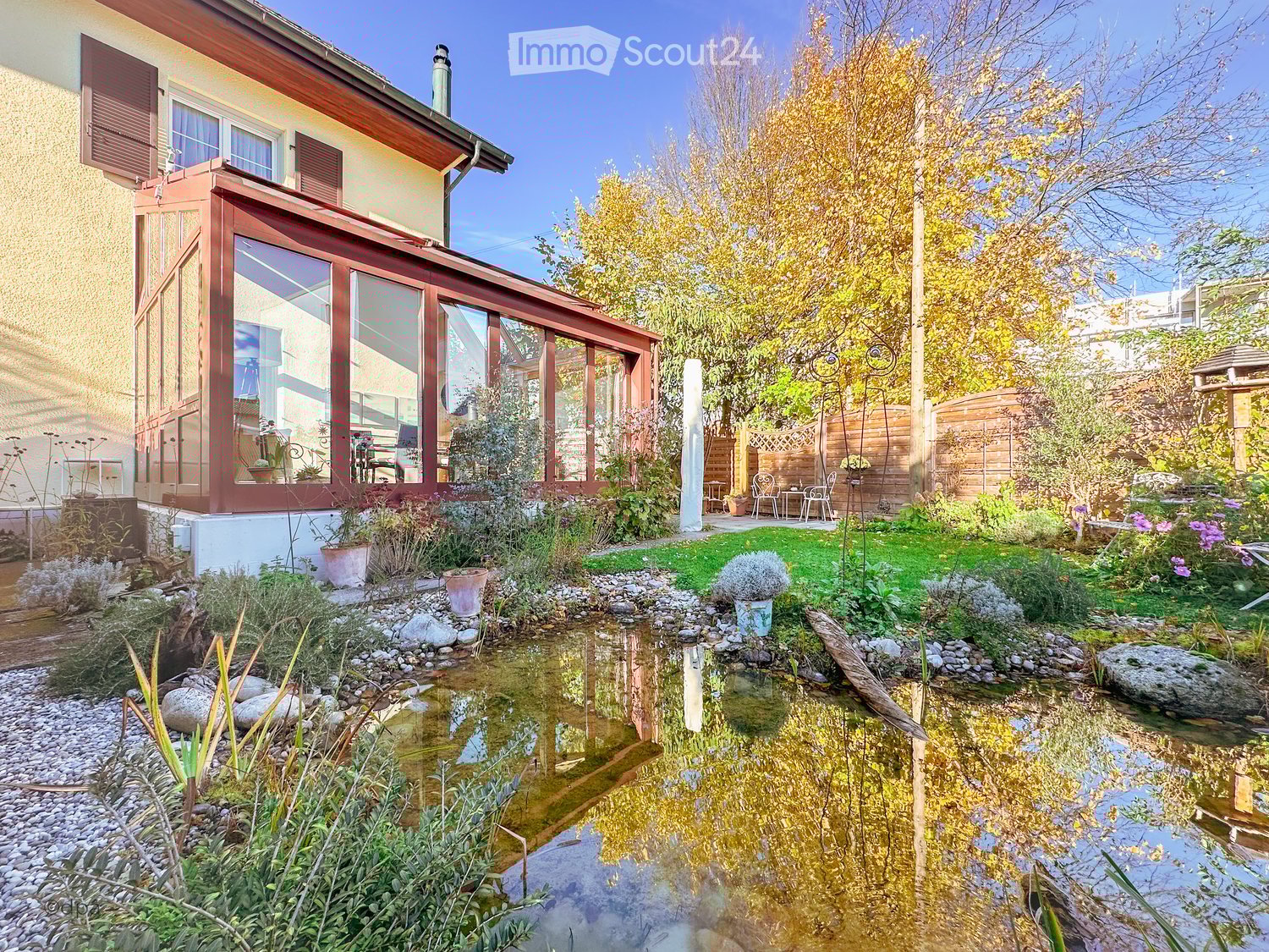 Pond with rocks, plants, and small bridge. Glass and wood conservatory with plants. Two tables and chairs. Trees and shrubs. Outdoor seating.