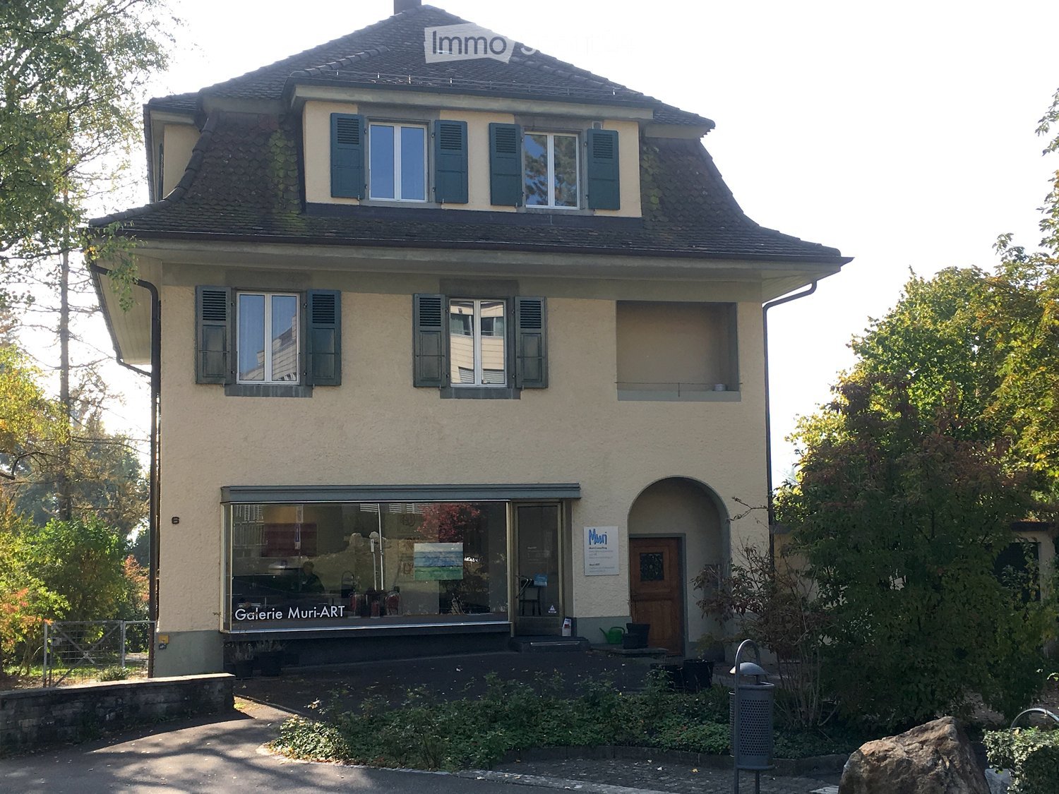 Two story house, beige walls, black roof, multiple windows, entrance with glass door, front garden, sign for Gallery