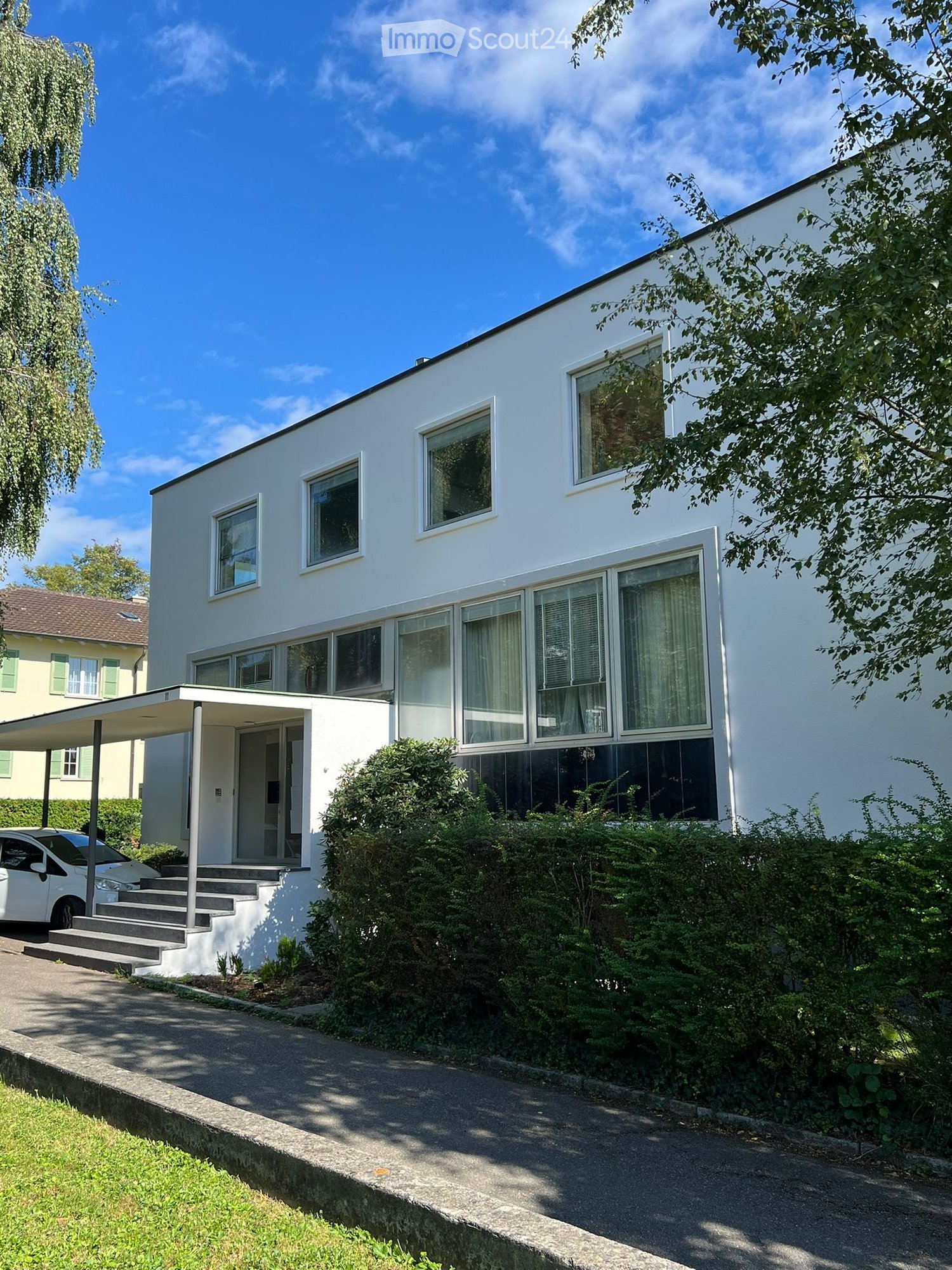 Modern white building with many windows, small bush, paved road, parking lot, car parked in front