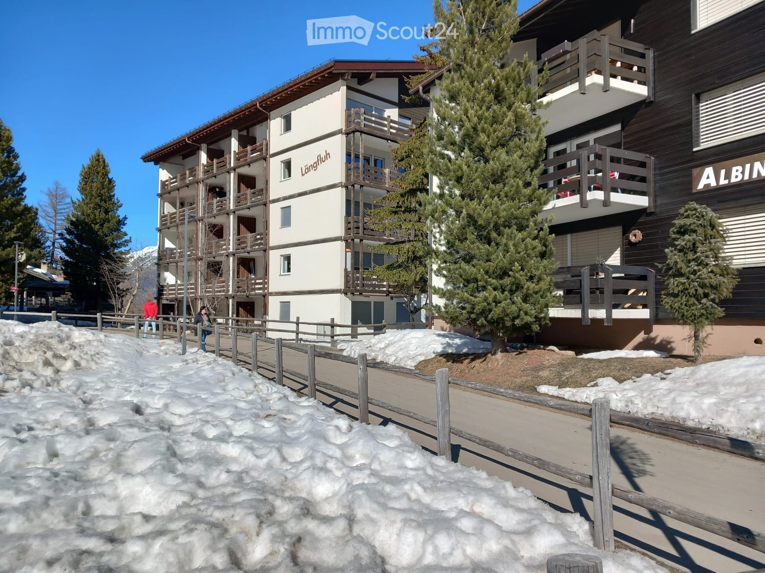 A two-story residential building with brown and white paint, balconies, a sign that reads 'Langfluh', a neighboring building labeled 'ALBIN', snow covering the ground, and people walking in front.