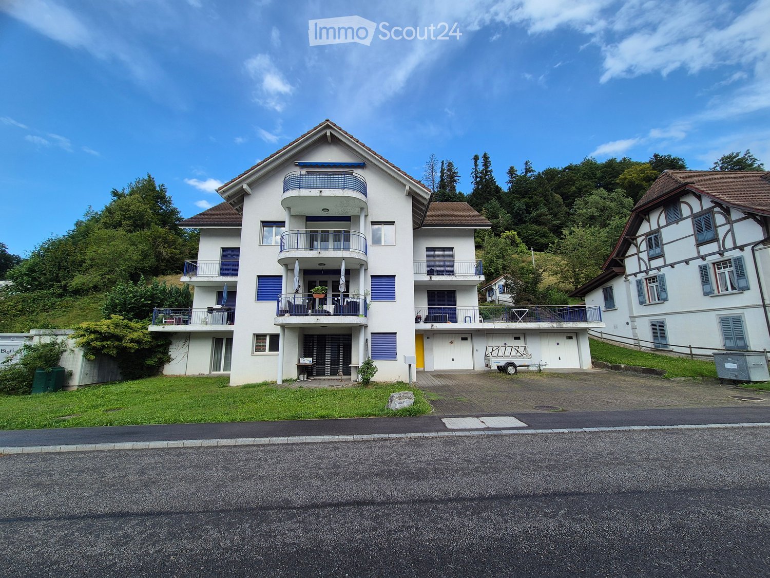Three-story apartment building, white exterior, brown roof, multiple balconies, entrance with stairs, fenced yard with green grass, adjacent driveway, another building in the background, trees in the distance