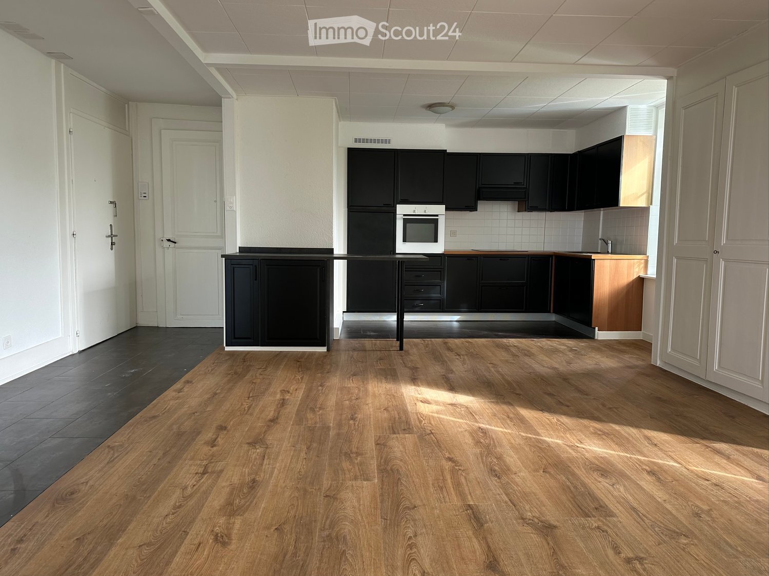 an open-plan living and dining area with a kitchen. The room has hardwood floors, a modern kitchen setup with white appliances, and dark wooden cabinets. The walls are painted white, and there is a wooden bar counter and a faucet mounted on the wall near