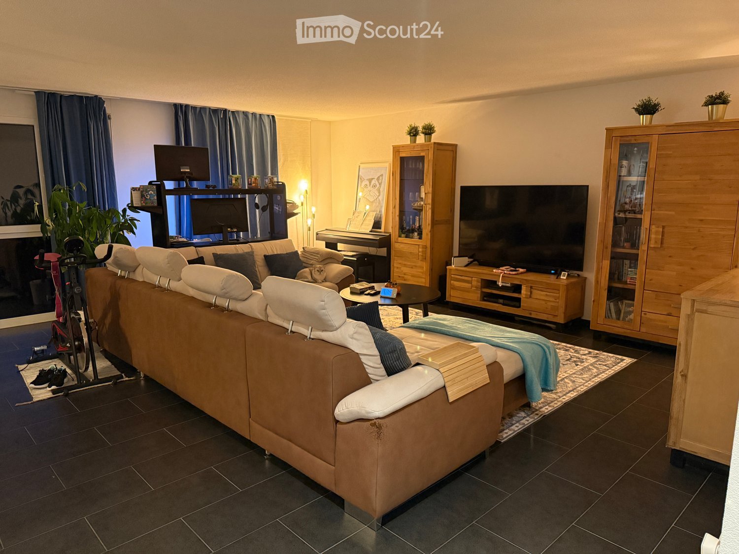 A living room with a large brown sofa, glass center table, a piano, a television, cabinets, potted plants, and a carpet.
