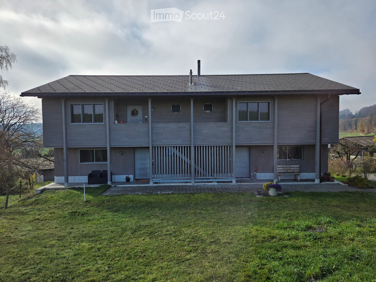 two-story house, gray exterior, large windows, raised ground floor, spacious front yard with grass, small pathway leading to the entrance, front seating area with two chairs, potted plant on the left, bench in front of one of the doors