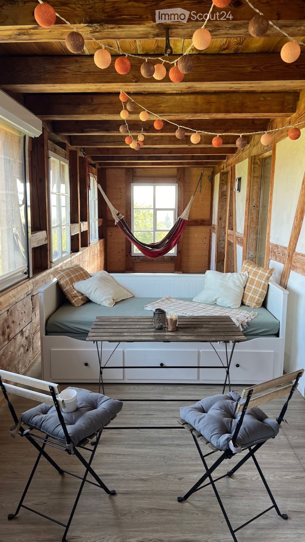 A cozy terrace space with a hanging hammock, daybed with drawers, wooden desk, two cushioned chairs, and decorative string lights on the ceiling.