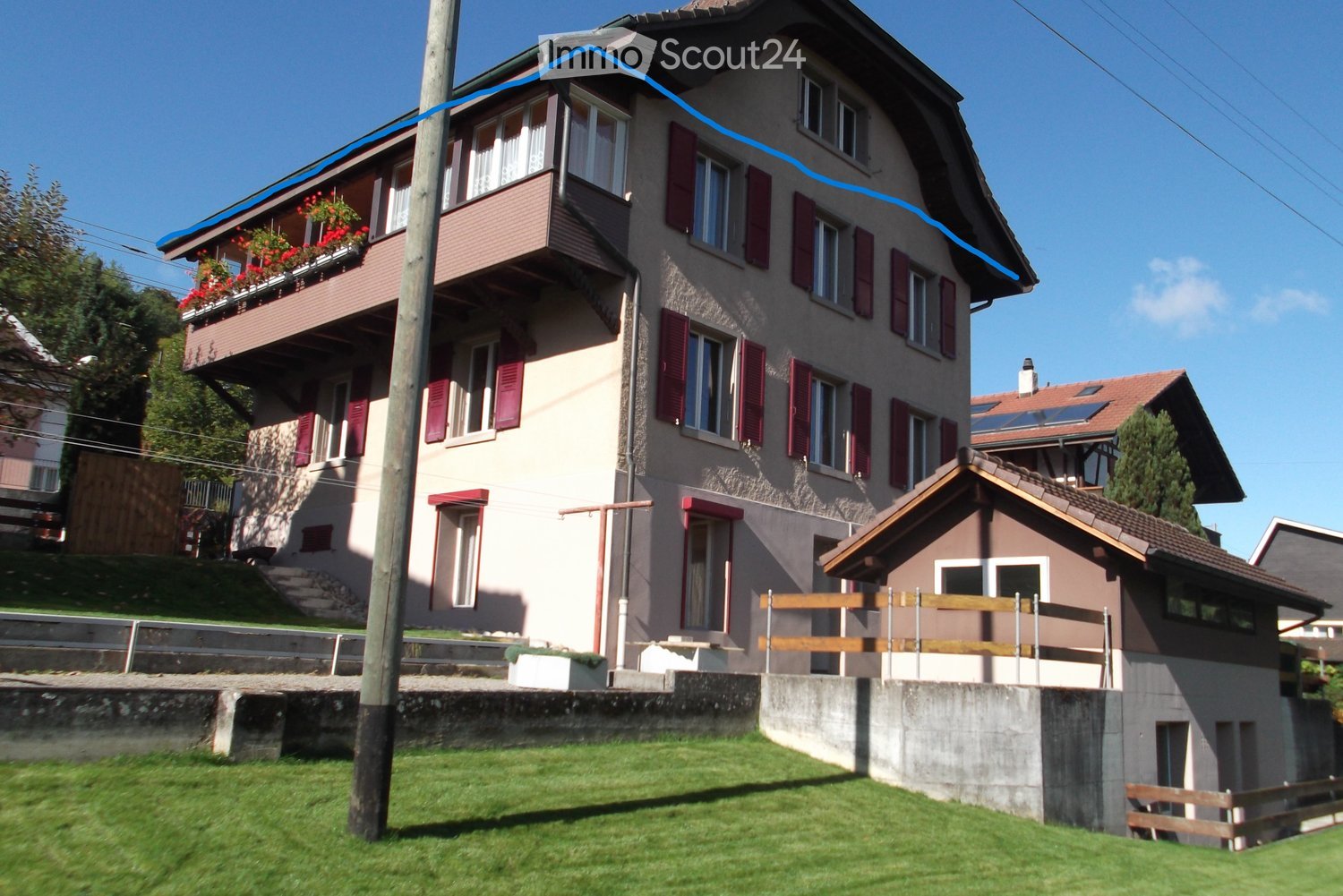 3-story residential building with red-tiled roof, brown exterior walls, and balconies with flower boxes. The building has a raised ground floor and is surrounded by a grassy area.