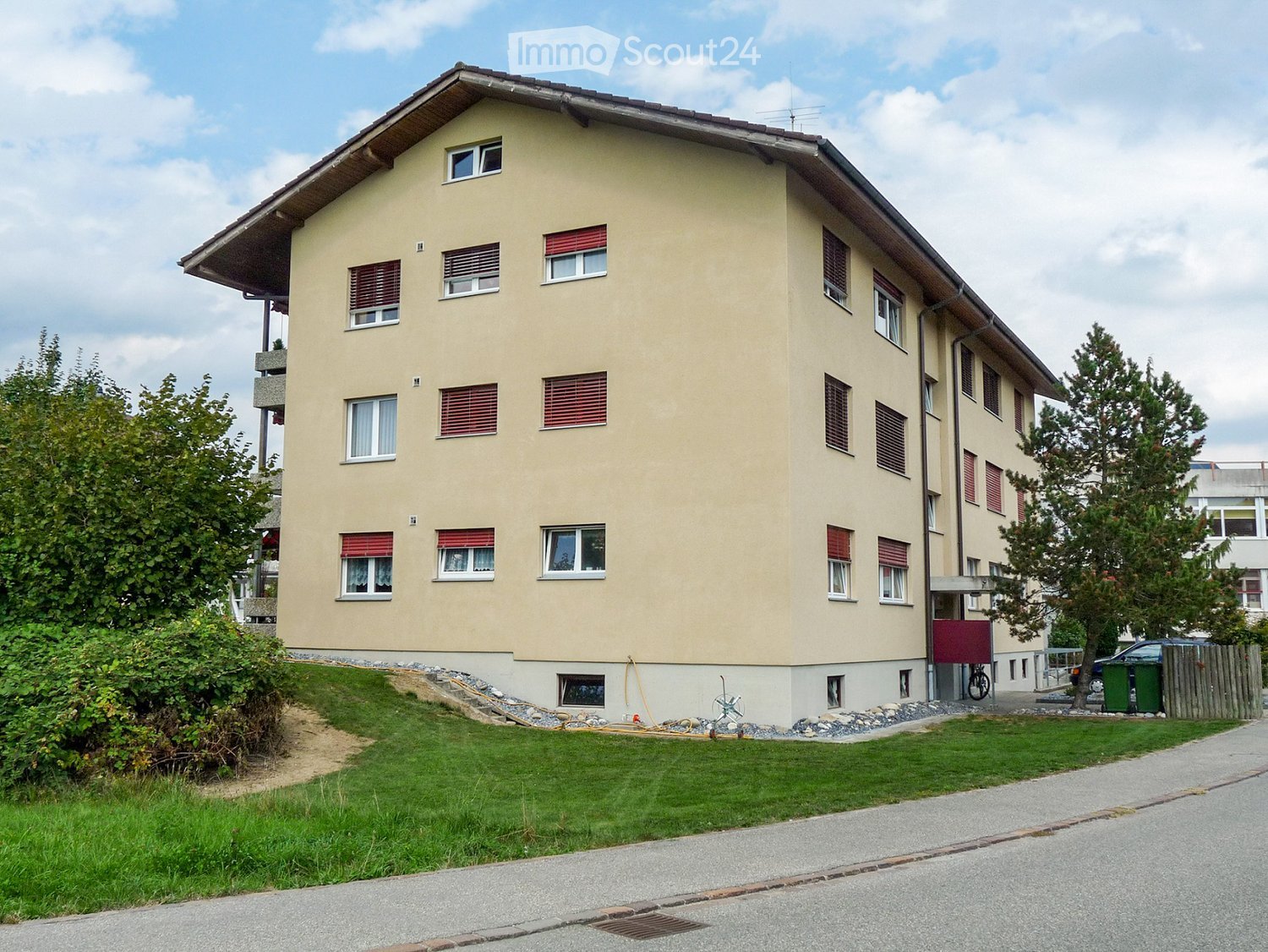 Three-story beige building with several balconies, fenced by shrubs, with a car parked nearby