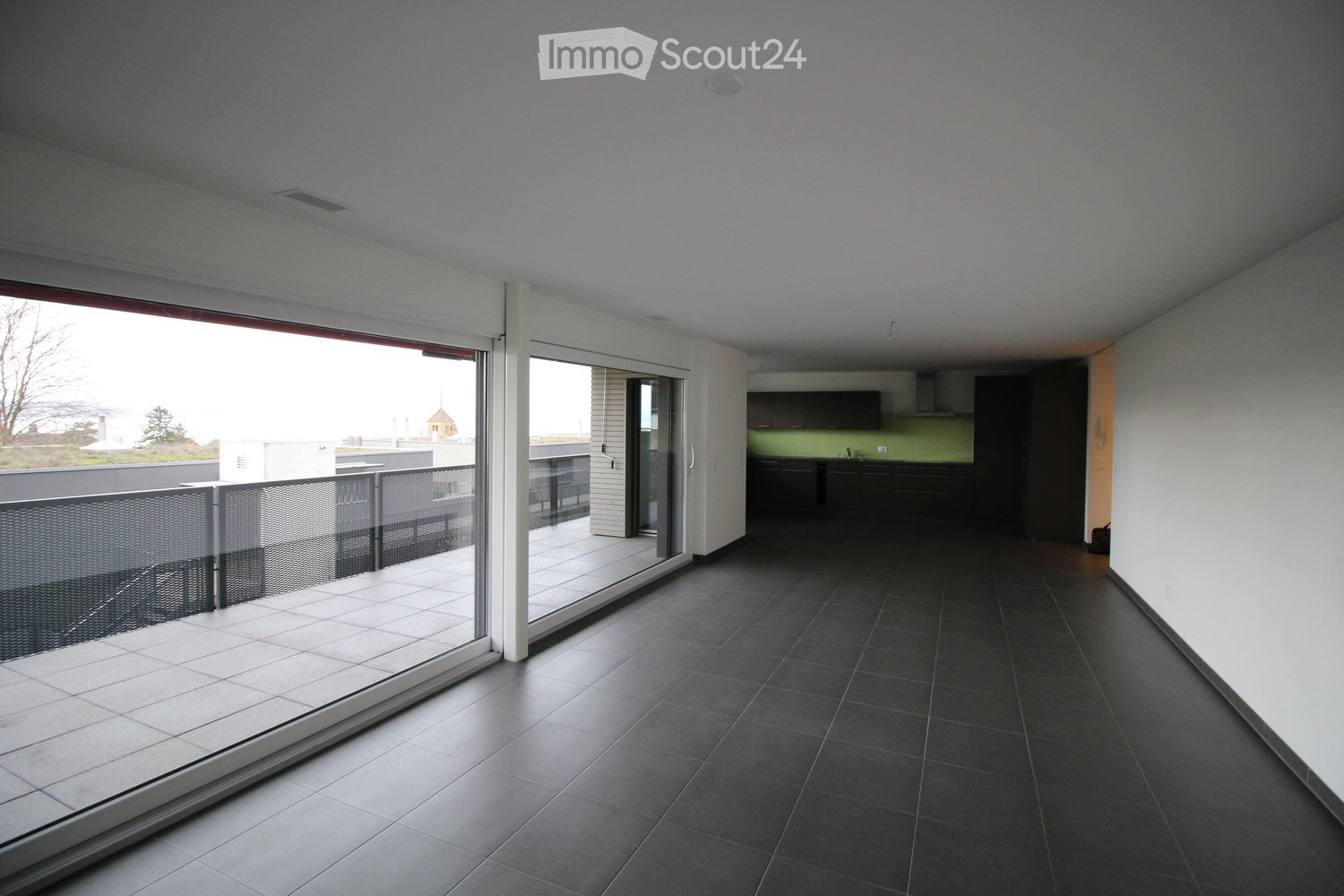 A large, empty room with gray tiles, white walls, glass doors leading to a balcony, kitchen, and black railings