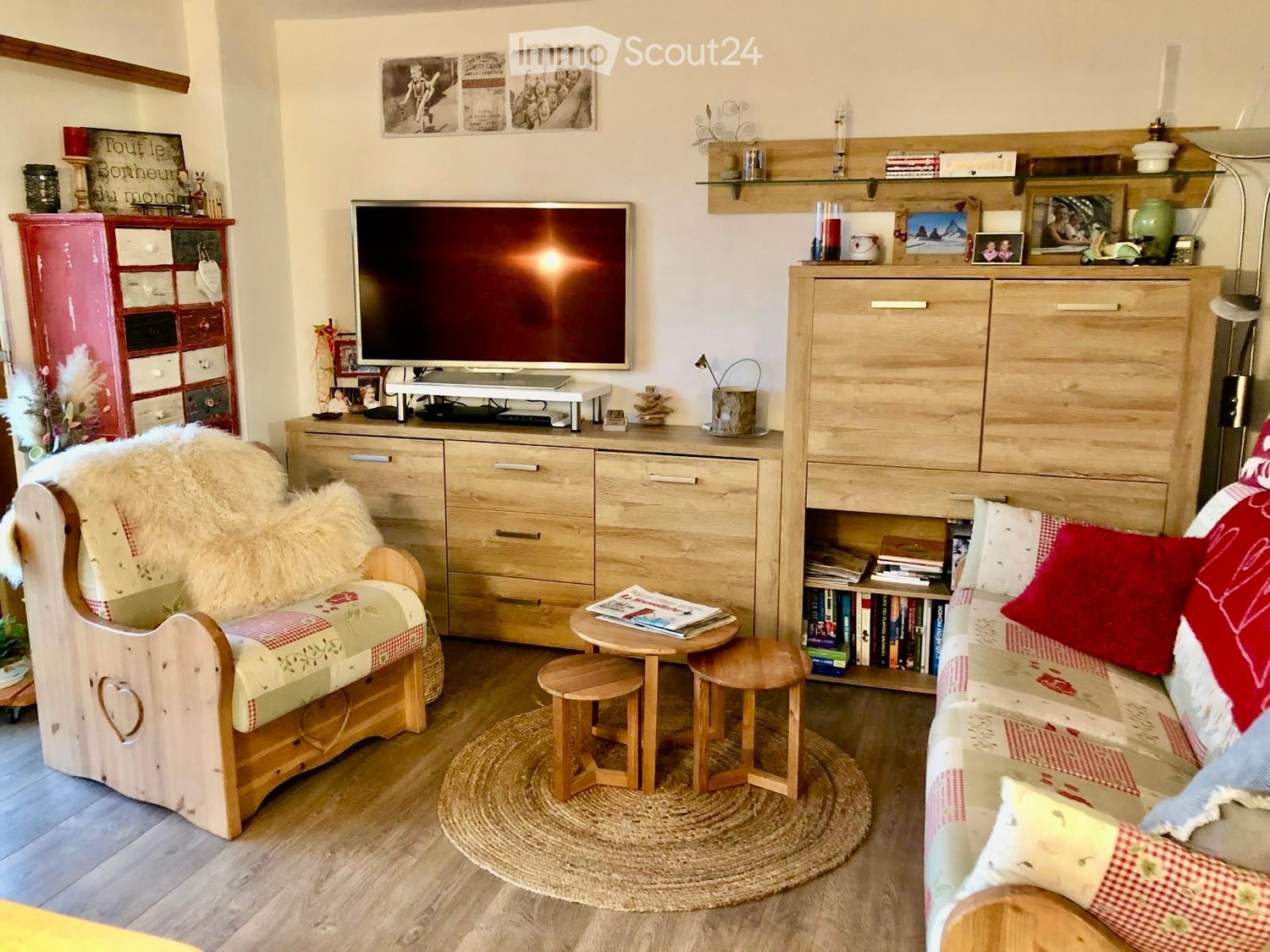 large wooden cabinet with drawers, TV on top, small round table with magazines, two small stools, sofa with pillows, wooden chair with fur, round rug on the floor, wall decorations and bookshelves, red shelving unit with various items