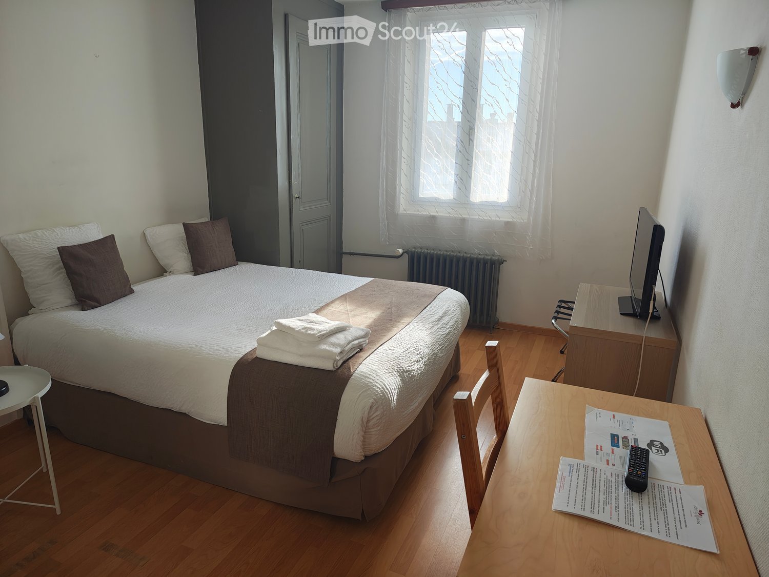 Double bed with pillows, white and brown bed sheets, wooden nightstand with TV and remote, radiator, folded wooden chair, wooden desk with papers