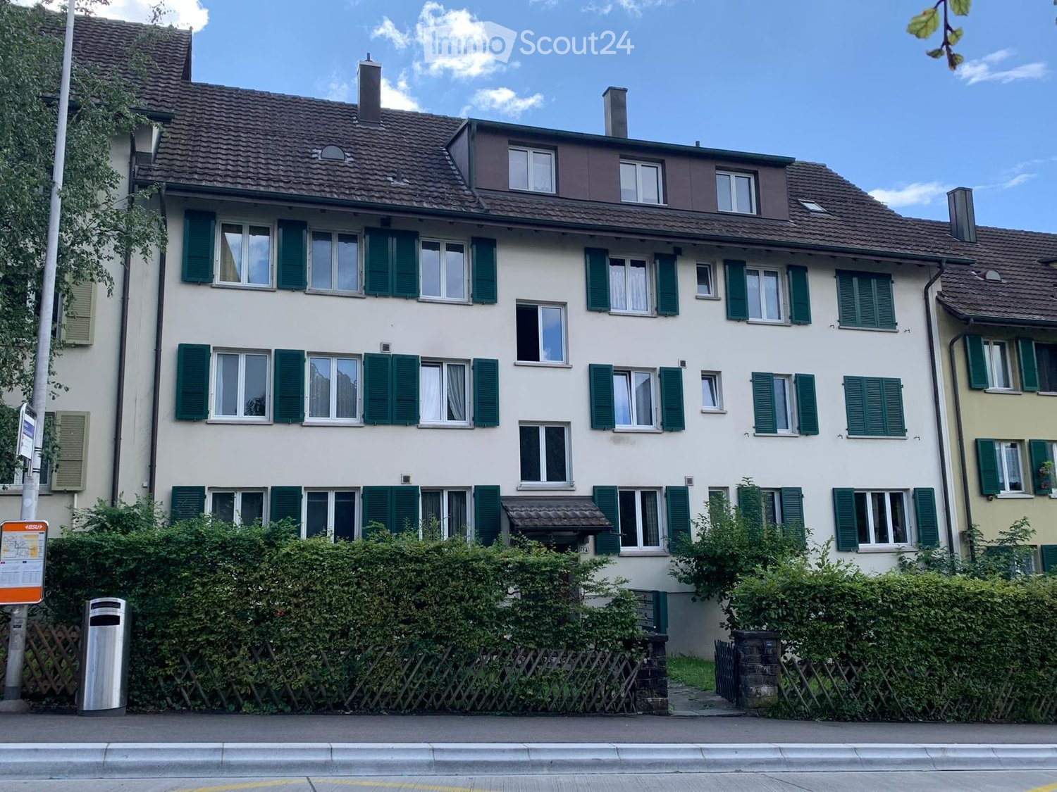 Multistory residential building, beige and green exterior, many windows, small shrubs, wooden fence, trash bin