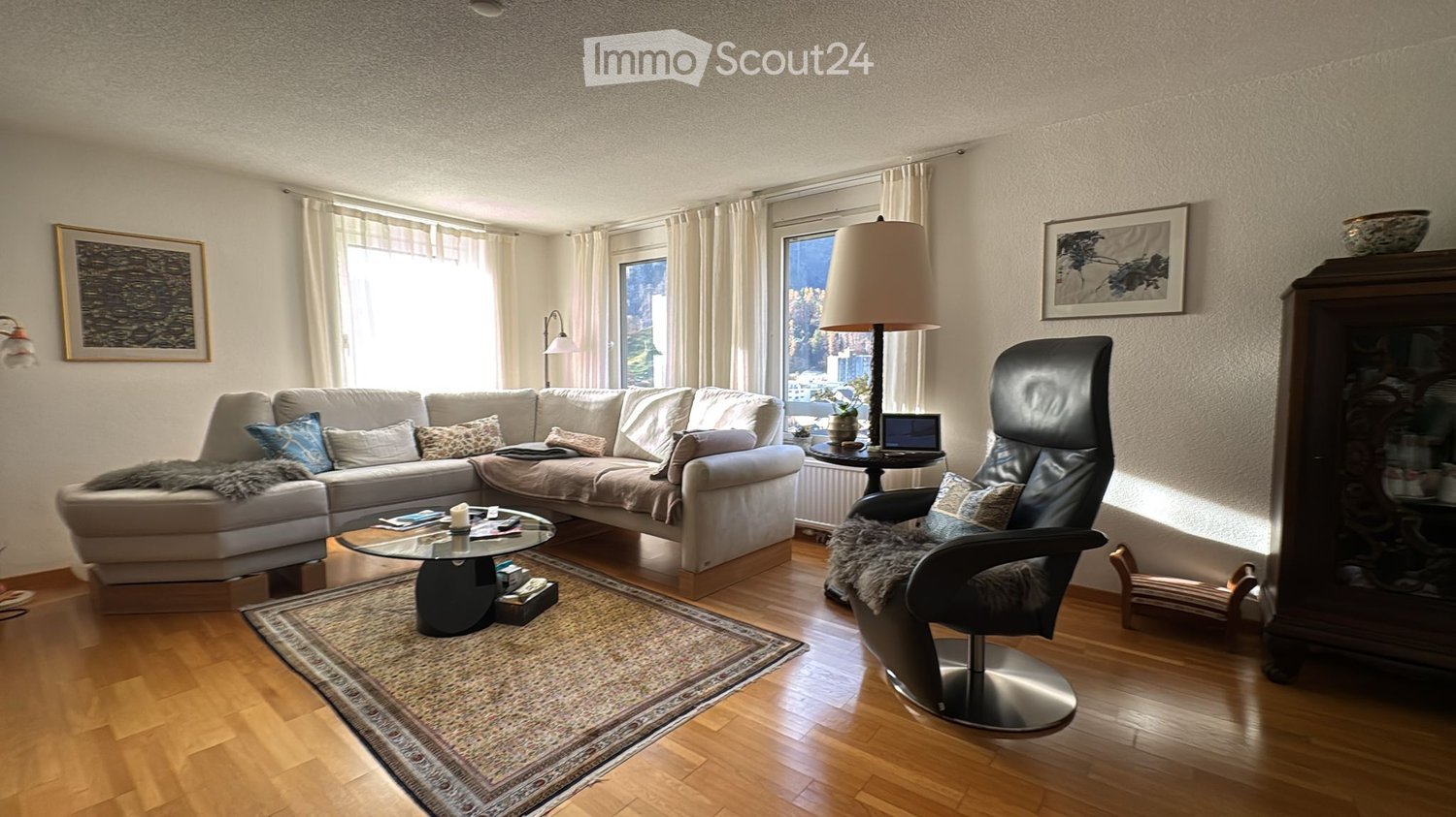 A U-shaped sofa, with a coffee table and rugs, a black chair with footrest, a table with a laptop on it, white walls and wooden floor.