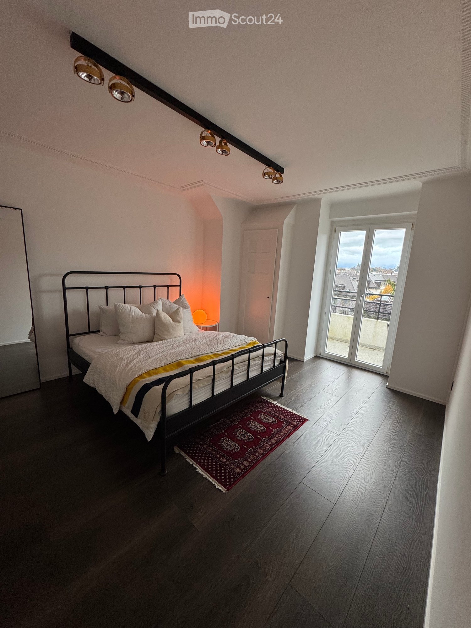 Bedroom with dark hardwood floor, black metal bed, sliding glass door leading to balcony, large windows, track lighting, mirror, and a rug.