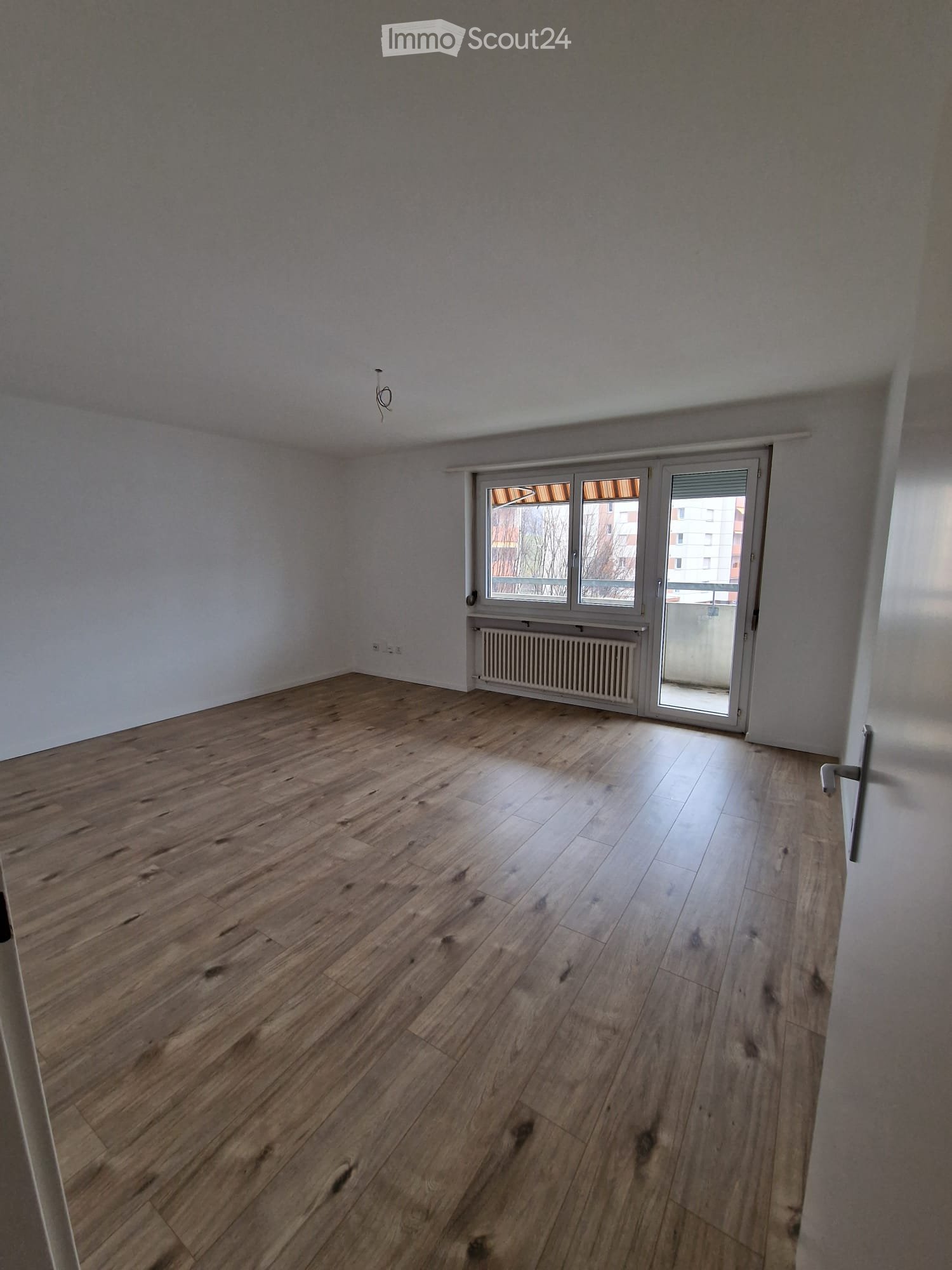 large empty room, hardwood floors, sliding glass door leading to balcony, white walls, window with a view to the outside