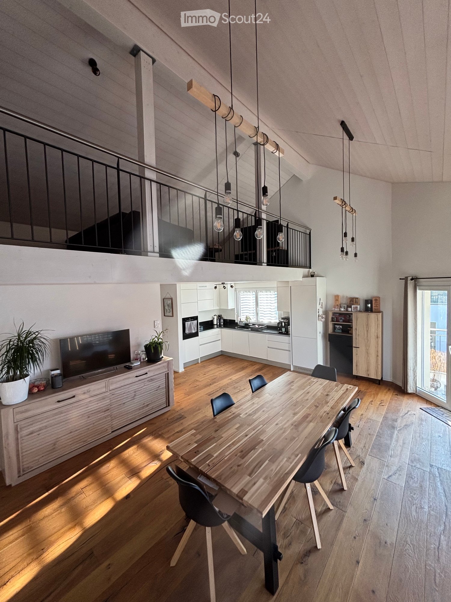 Open concept living room and kitchen with wooden flooring and white walls. Dining table with chairs in the living room. TV and potted plant on a wooden dresser next to the couch. Modern kitchen with appliances.