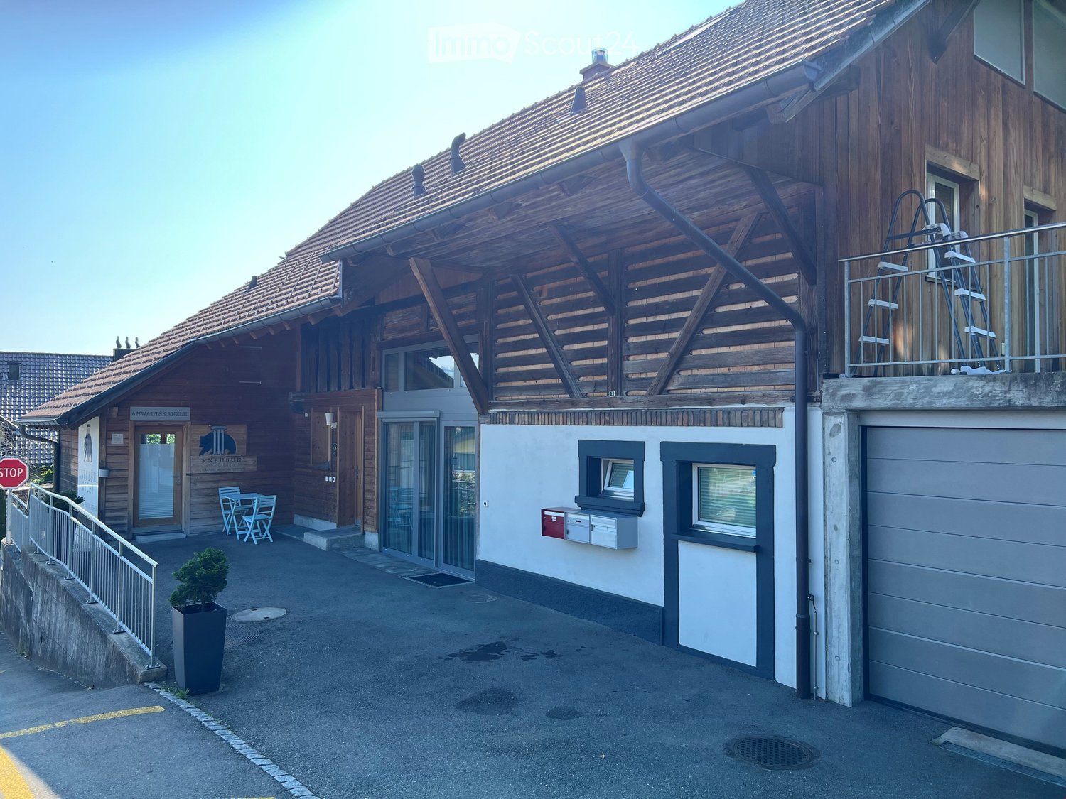 two-story building with wooden exterior, large garage with ladder, stairs leading to an upper floor balcony, small garden area with potted plant