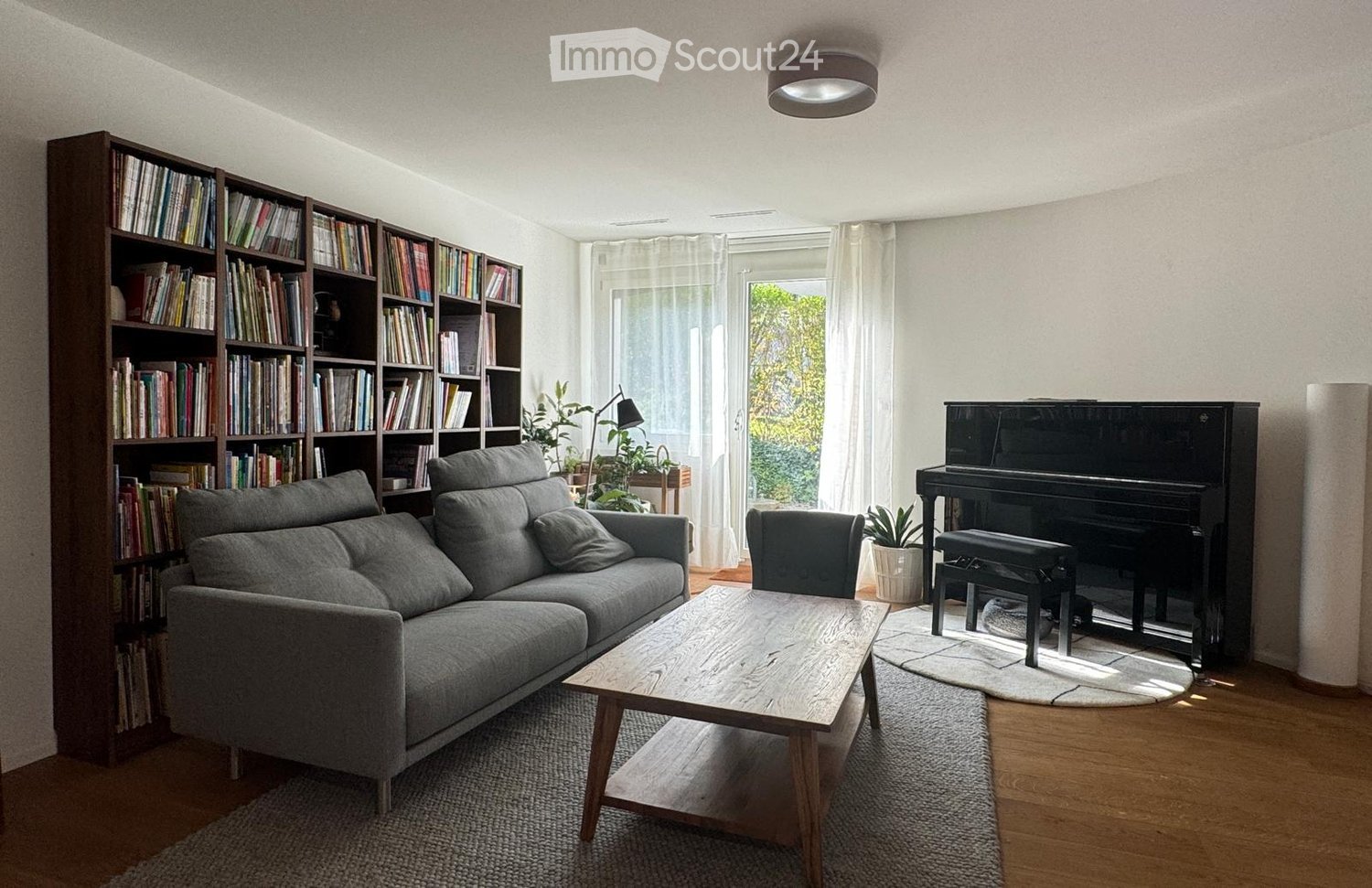 Wooden coffee table, gray couch, bookshelf filled with books, black piano, potted plants